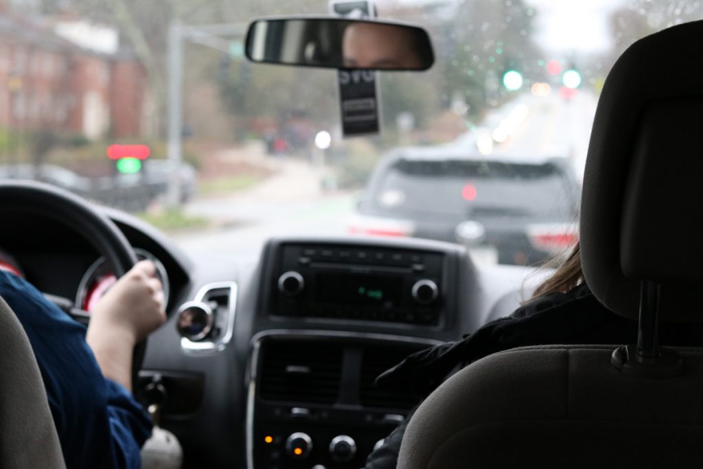 A driver and passenger sit in the front of a paratransit van with the driver's face visible in the rearview mirror.