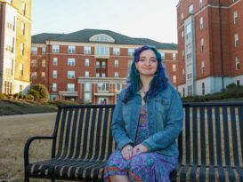 Photo of Mary Jo Eden a University of Georgia student with purple and teal hair to match her colorful dress, sitting on a bench in a courtyard surrounded by dorm buildings.