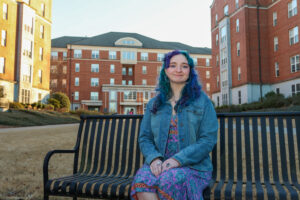 Photo of Mary Jo Eden a University of Georgia student with purple and teal hair to match her colorful dress, sitting on a bench in a courtyard surrounded by dorm buildings.