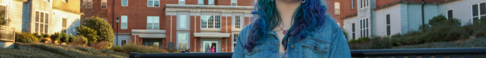 Photo of Mary Jo Eden a University of Georgia student with purple and teal hair to match her colorful dress, sitting on a bench in a courtyard surrounded by dorm buildings.