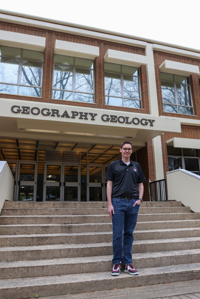 Dr. Christopher Elcik stands with one hand in his pocket on the steps of the geography geology building on UGA campus. 