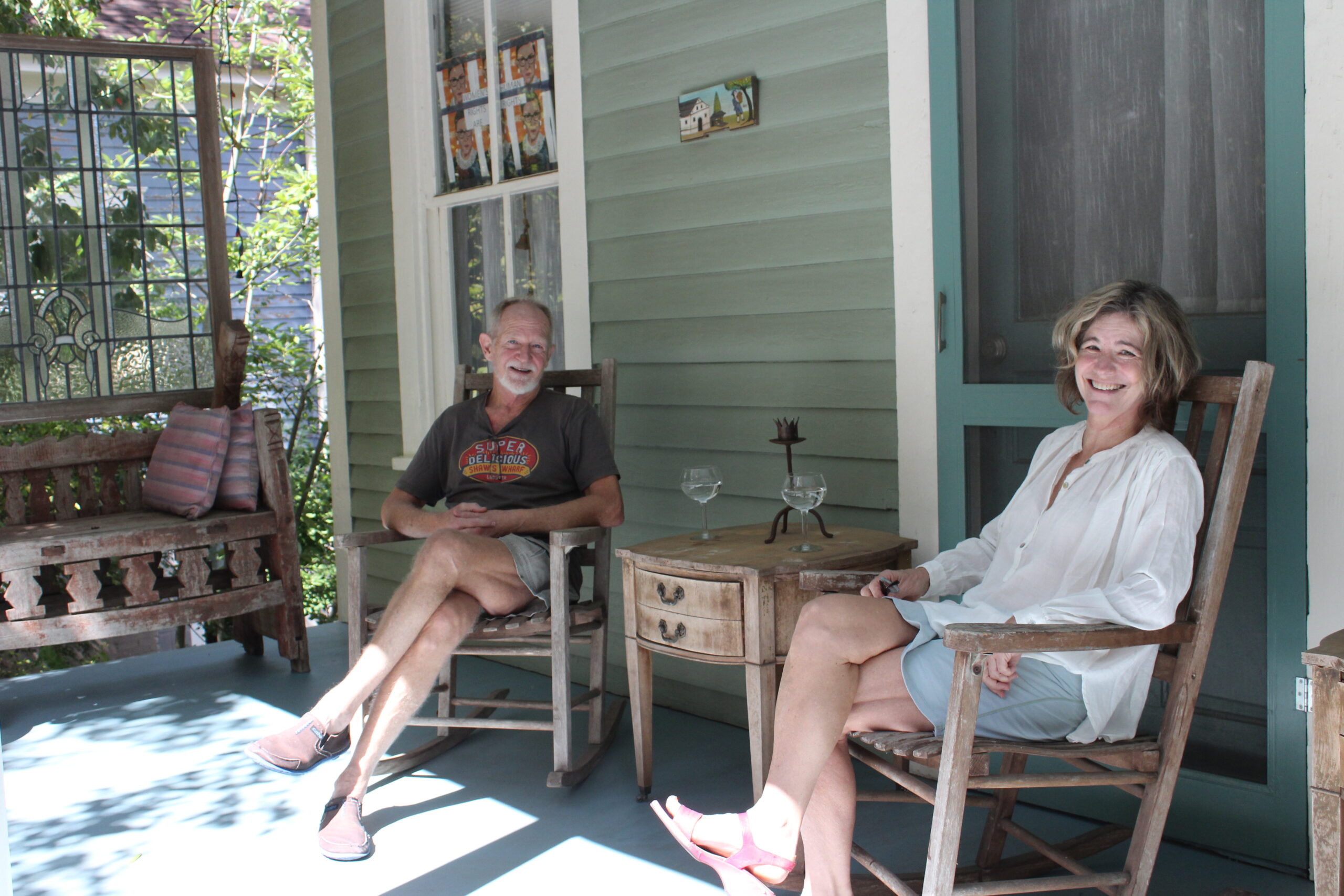 Two people sit in chairs on a porch, smiling at the camera.