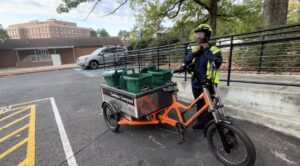 A UGA Campus Compost intern stands next to a bright orange cargo bike loaded with green compost bins labeled "Campus Compost." The intern wears a reflective jacket and a helmet, adjusting the chin strap.