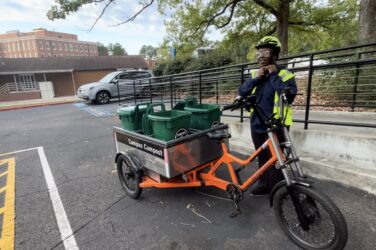 A UGA Campus Compost intern stands next to a bright orange cargo bike loaded with green compost bins labeled "Campus Compost." The intern wears a reflective jacket and a helmet, adjusting the chin strap.