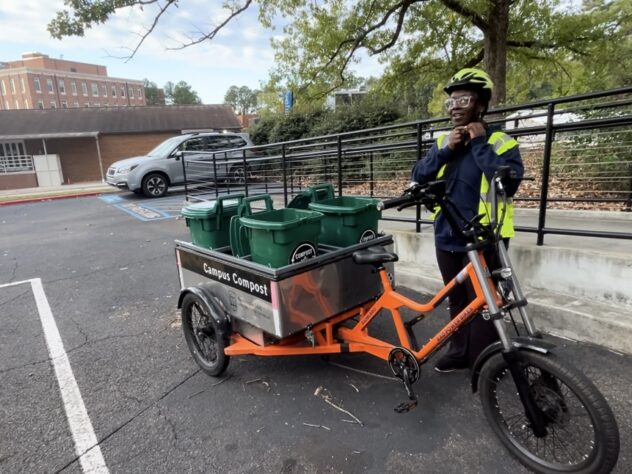 A UGA Campus Compost intern stands next to a bright orange cargo bike loaded with green compost bins labeled "Campus Compost." The intern wears a reflective jacket and a helmet, adjusting the chin strap.