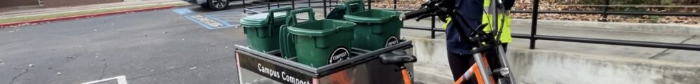 A UGA Campus Compost intern stands next to a bright orange cargo bike loaded with green compost bins labeled "Campus Compost." The intern wears a reflective jacket and a helmet, adjusting the chin strap.