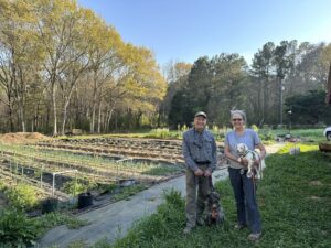 A man and a woman stand with two dogs in front of community food plots.