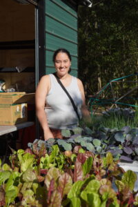 A smiling person in a white tank top stands behind a table of fresh leafy vegetables beside a green metal building, with trees and sunlight in the background.