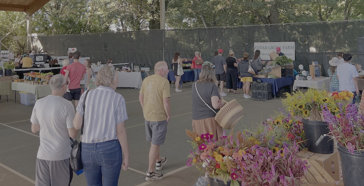 People browse an outdoor market, with vibrant flower bouquets in the foreground and bustling market stalls in the background, all set beneath a canopy of trees with sunlight streaming through.