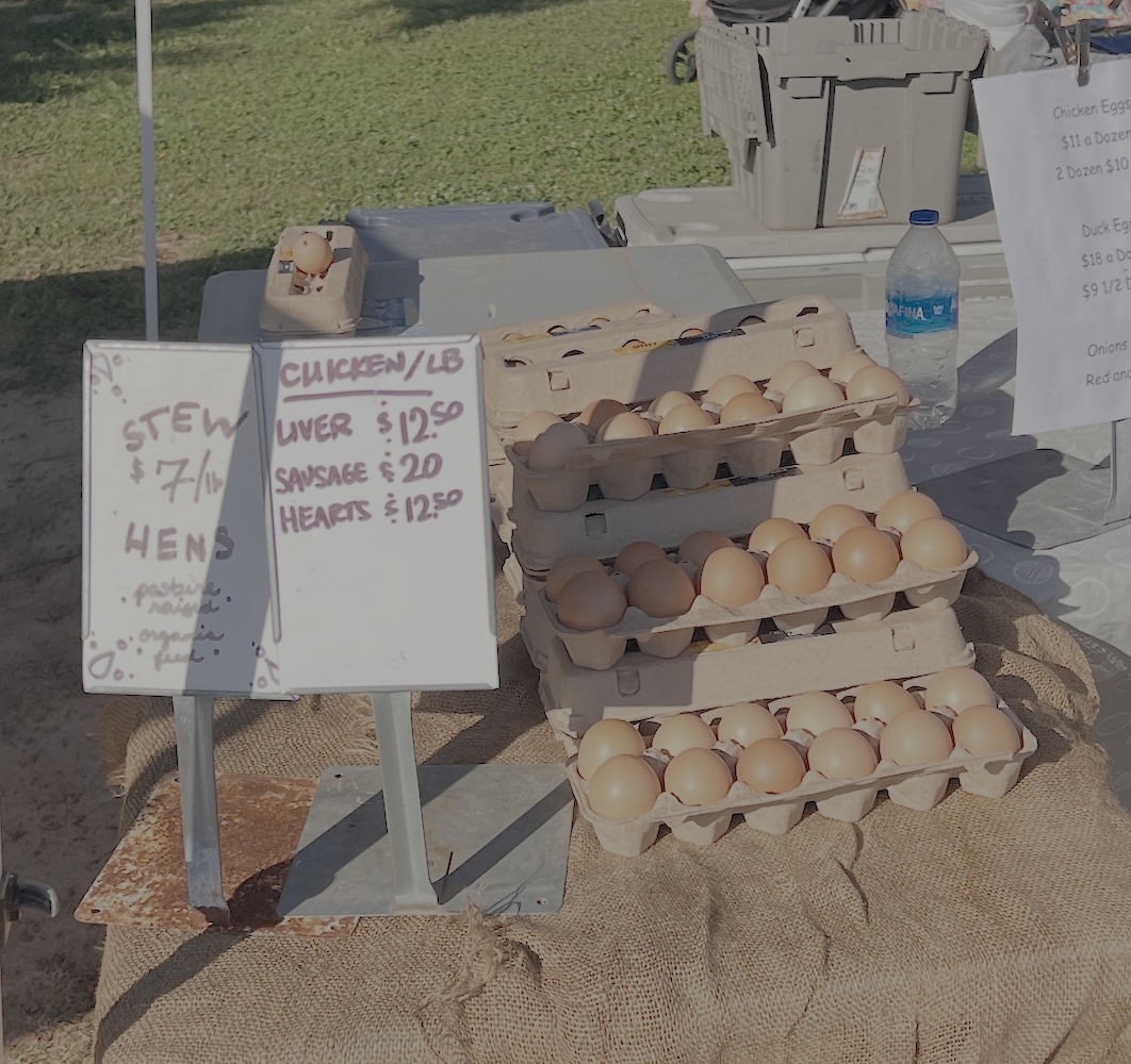 Cartons of brown eggs are stacked on a burlap-covered table at an outdoor market. A handwritten sign lists chicken products and prices, while a water bottle, baskets, and green grass complete the lively market scene.