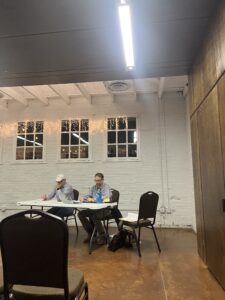 Two men sit at a folding table in a room with white brick walls and string lights. Papers and laptops are on the table. Empty chairs face the table, and the floor is brown. The ceiling has exposed beams and bright lighting.