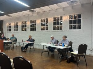 Five people sit at tables arranged in a row in a bright room with white brick walls, windows, and string lights. Papers, laptops, and drinks are on the tables, suggesting a meeting or discussion is taking place.