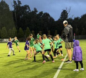 Young girls in teal and purple jerseys play soccer on a green field as a coach and a child in a purple raincoat watch nearby. The scene is lively, with trees and a rainy sky in the background.