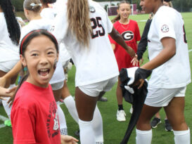 A young girl in a red shirt smiles excitedly while giving high-fives to female soccer players in white uniforms on a field. Other players and children are visible in the background.