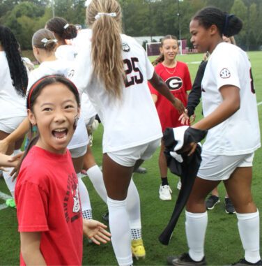 A young girl in a red shirt smiles excitedly while giving high-fives to female soccer players in white uniforms on a field. Other players and children are visible in the background.