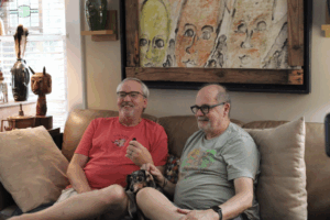 Two older men sit close together on a beige couch, smiling and holding hands, with a small dog on their laps. Colorful abstract art and decorative items are displayed in the cozy, well-lit living room.