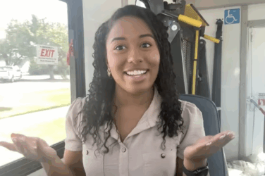 A reporter on a bus, smiling with both hands turned up to show options.