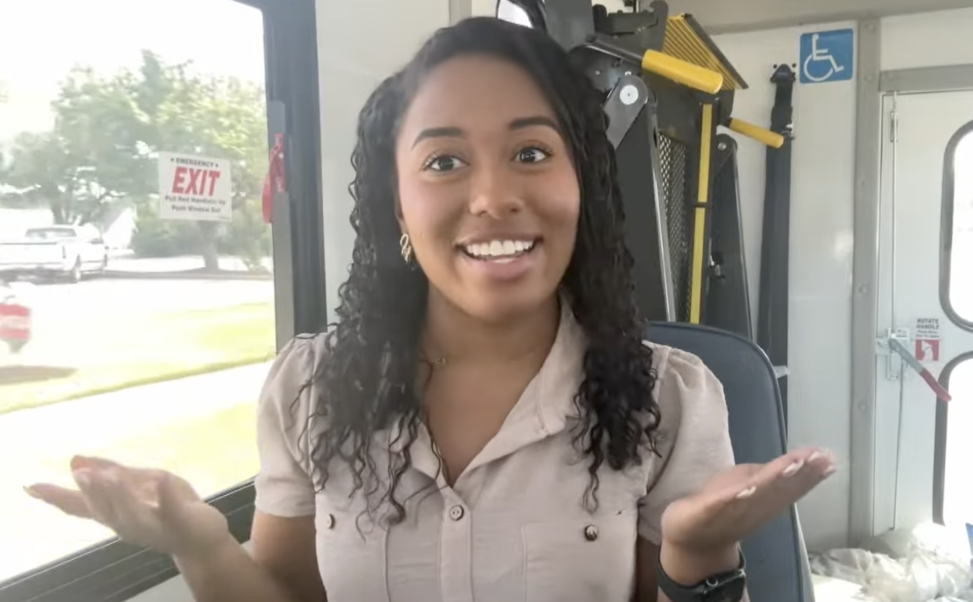 A reporter on a bus, smiling with both hands turned up to show options.