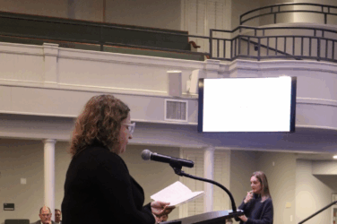 Woman stands at a podium and speaks into a microphone at a board of education meeting