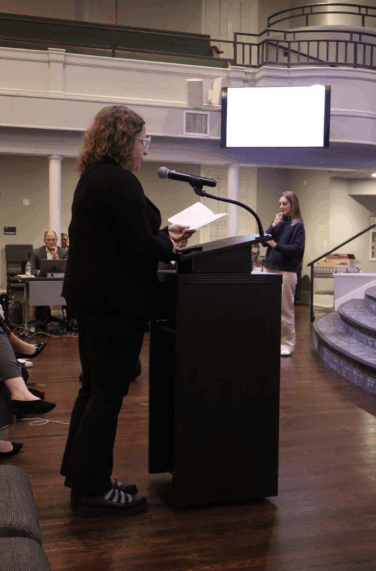 Woman stands at a podium and speaks into a microphone at a board of education meeting