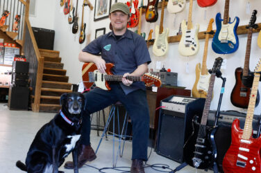 A man smiles on a stool with a guitar hands and a black lab dog at his side.