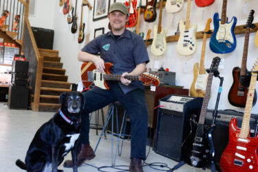 A man smiles on a stool with a guitar hands and a black lab dog at his side.