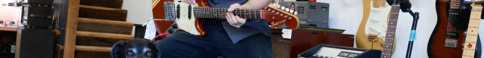 A man smiles on a stool with a guitar hands and a black lab dog at his side.