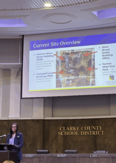 Woman speaks at podium during meeting with projection behind her.