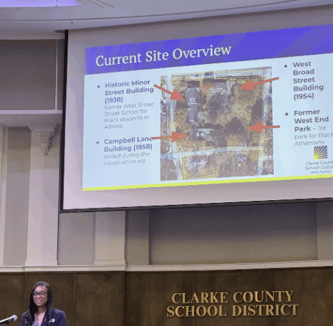 Woman speaks at podium during meeting with projection behind her.