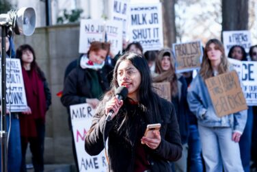 A woman speaks into a microphone with people protesting with signs in the background.