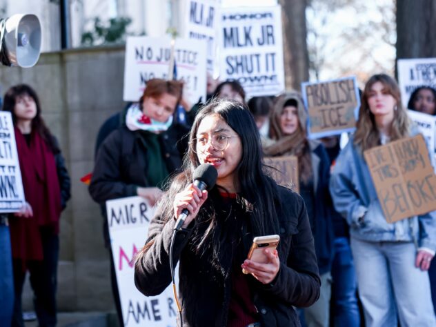 A woman speaks into a microphone with people protesting with signs in the background.