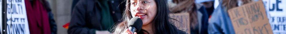 A woman speaks into a microphone with people protesting with signs in the background.