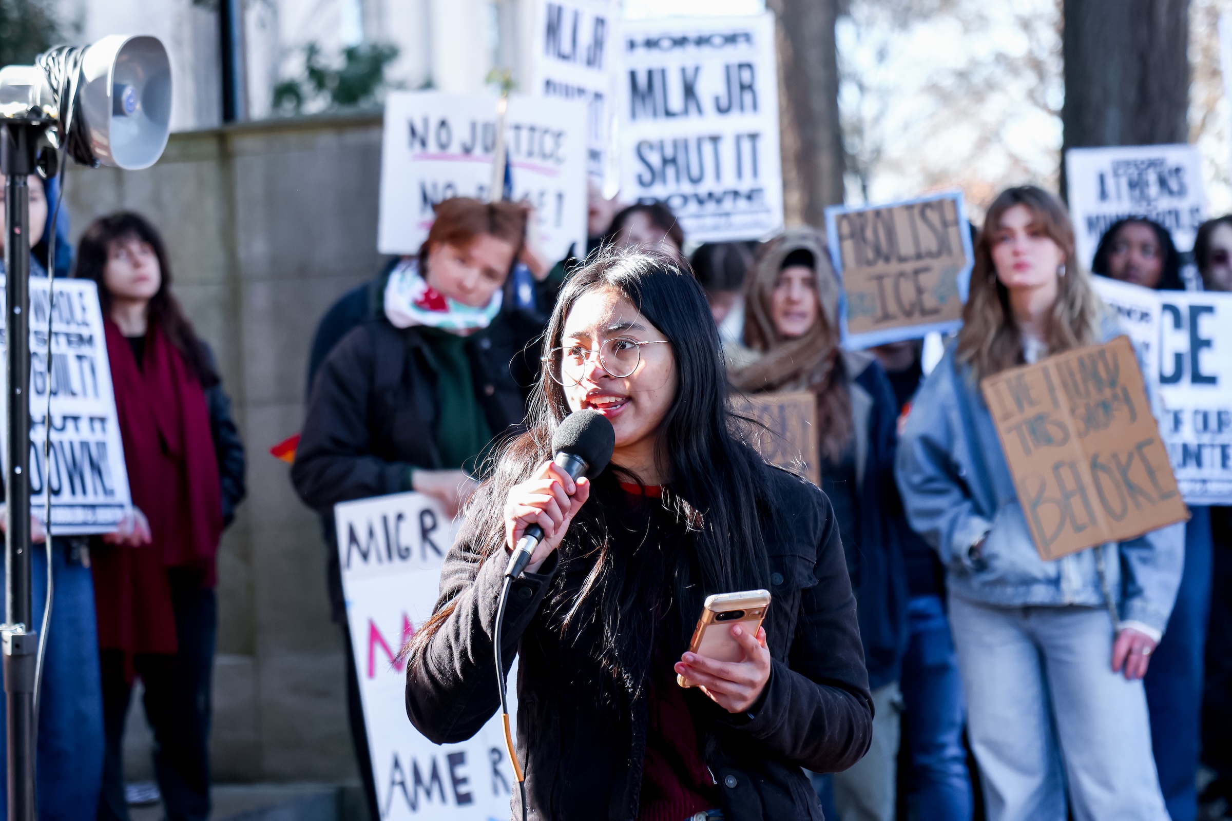 A woman speaks into a microphone with people protesting with signs in the background.