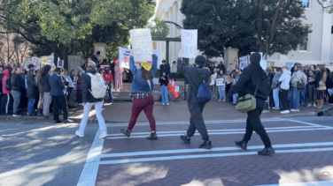 Protestors walk with signs across the screen in front of a college campus.