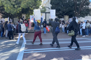 Protestors walk with signs across the screen in front of a college campus.
