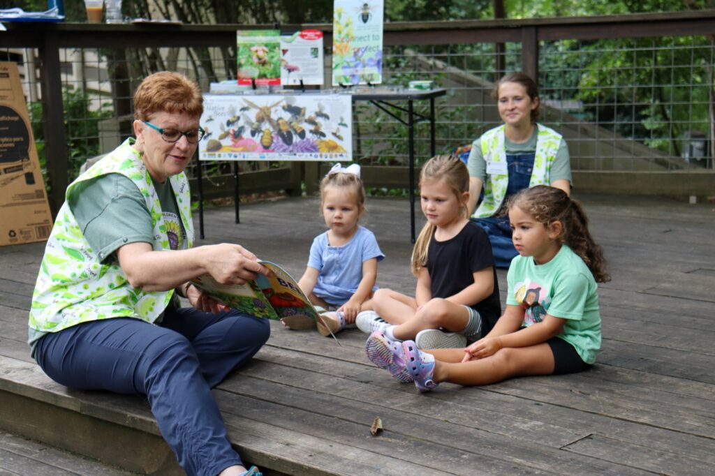A woman reads a book to three small children while sitting on a wooden platform in the woods.