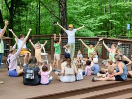A group of adults and children put their arms in the air while sitting on a platform in the woods.