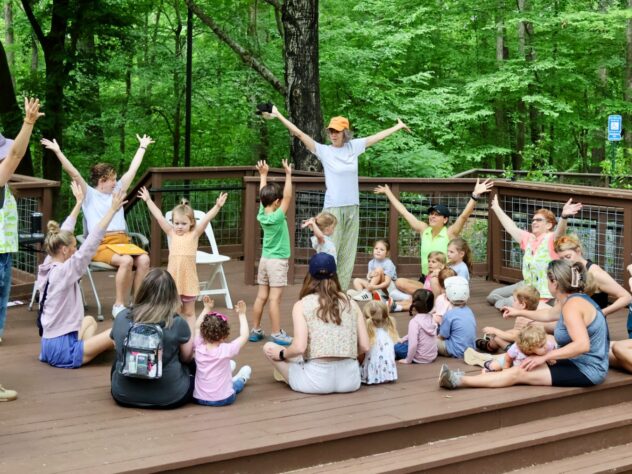 A group of adults and children put their arms in the air while sitting on a platform in the woods.