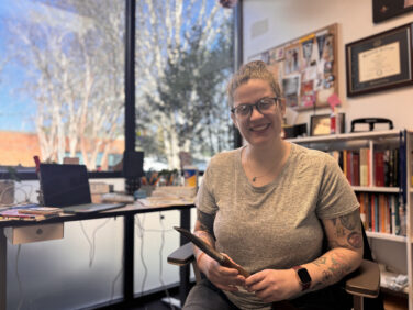 Woman sits with a garden tool in her hand at a desk and smiles at the camera