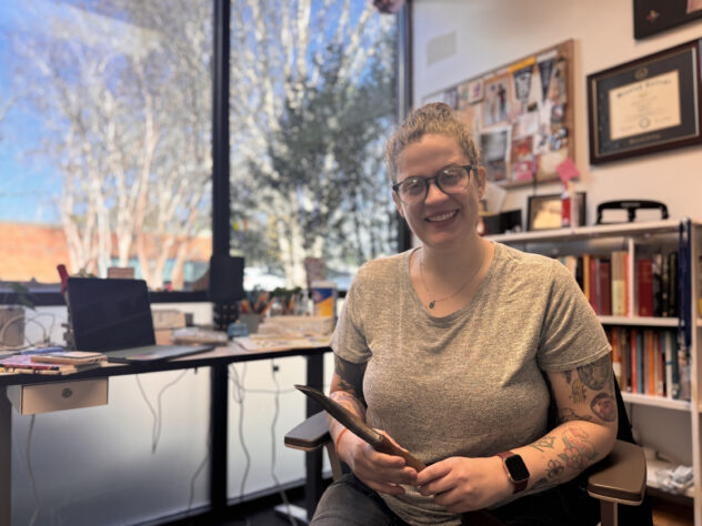 Woman sits with a garden tool in her hand at a desk and smiles at the camera