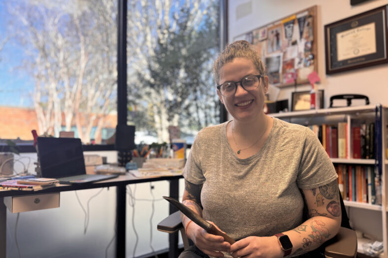 Woman sits with a garden tool in her hand at a desk and smiles at the camera