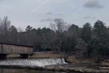 A covered wooden bridge spans a river beside a low dam, where water flows evenly over the edge into a calm pool below. Bare winter trees and evergreens line the banks under an overcast sky.
