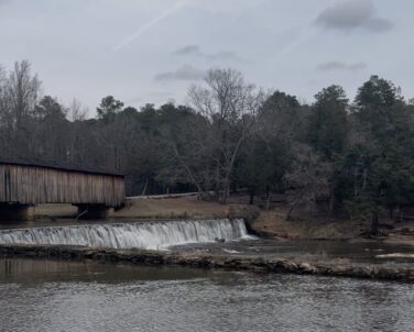 A covered wooden bridge spans a river beside a low dam, where water flows evenly over the edge into a calm pool below. Bare winter trees and evergreens line the banks under an overcast sky.