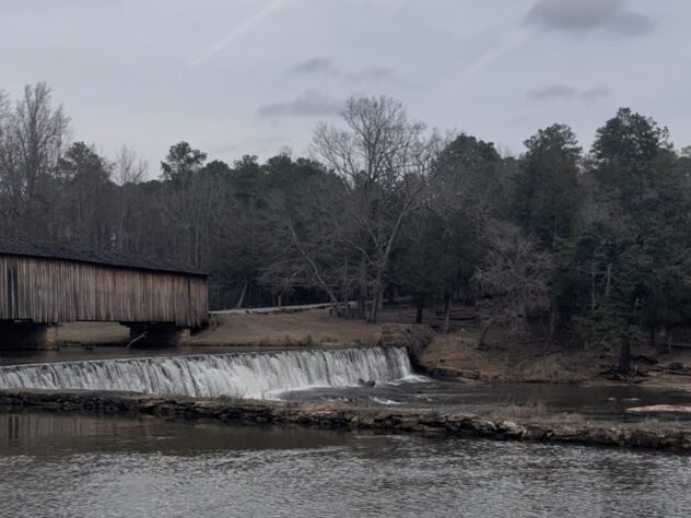A covered wooden bridge spans a river beside a low dam, where water flows evenly over the edge into a calm pool below. Bare winter trees and evergreens line the banks under an overcast sky.
