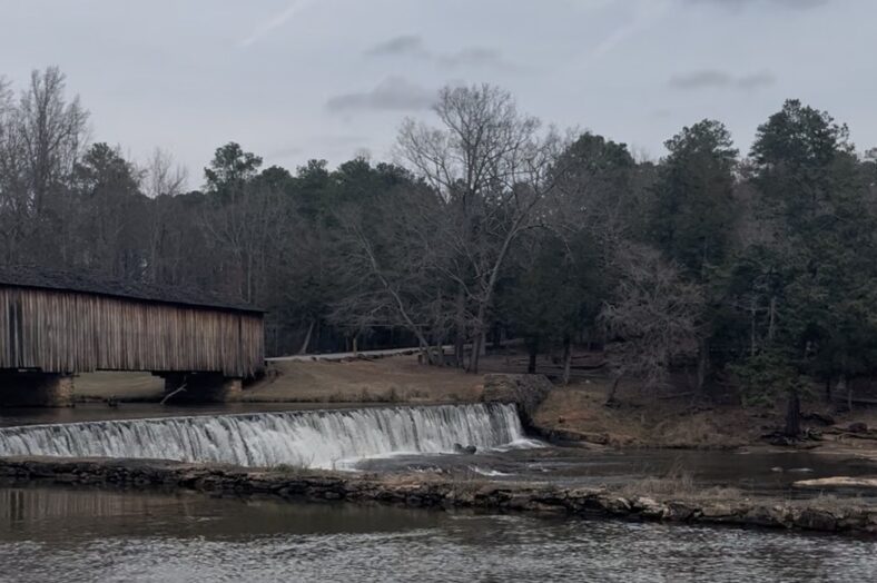 A covered wooden bridge spans a river beside a low dam, where water flows evenly over the edge into a calm pool below. Bare winter trees and evergreens line the banks under an overcast sky.
