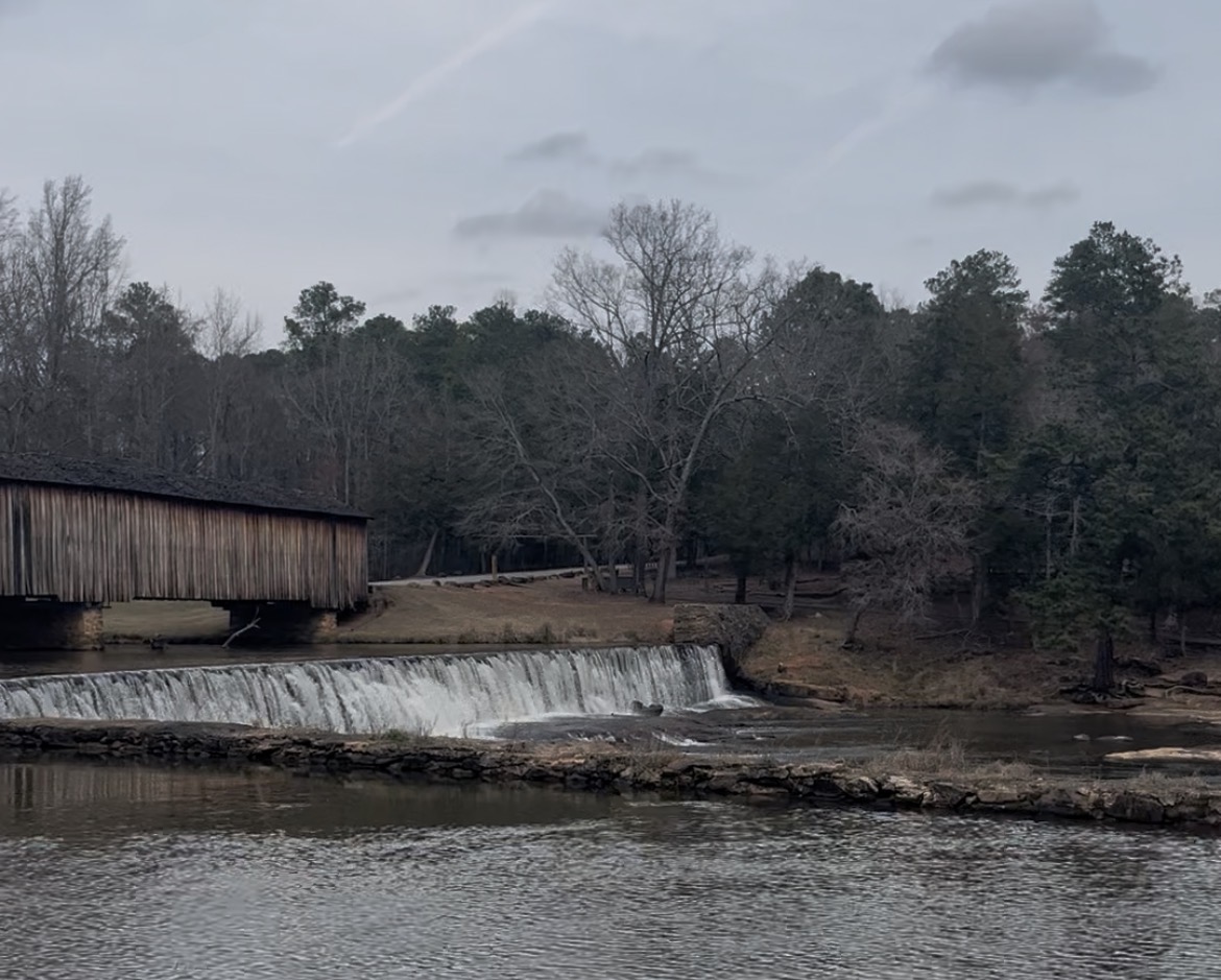 A covered wooden bridge spans a river beside a low dam, where water flows evenly over the edge into a calm pool below. Bare winter trees and evergreens line the banks under an overcast sky.