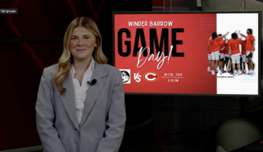 Sports journalist stands in front of a screen with game day logo