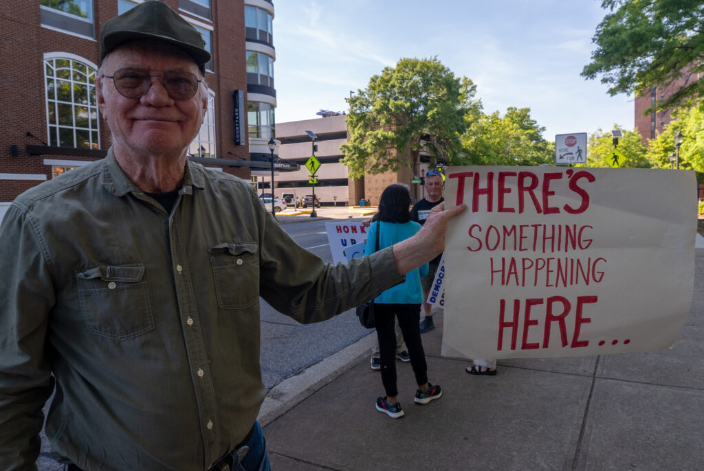An old man holds a sign in front of the camera