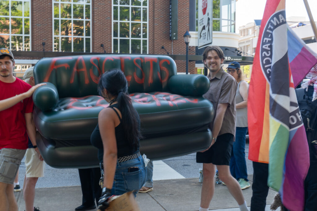 Two men walk holding a green inflatable couch spray painted with the word Fascists visable