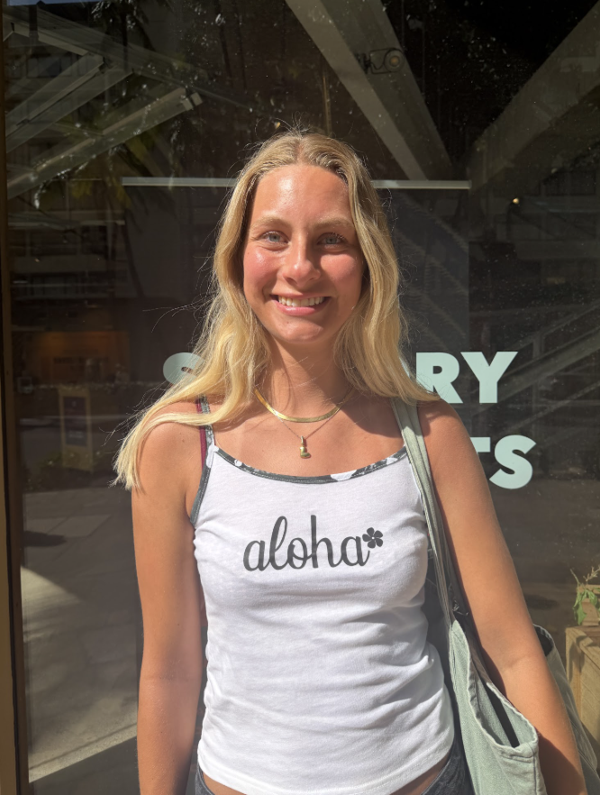 A woman smiles into the camera with a white "aloha" tank top on.
